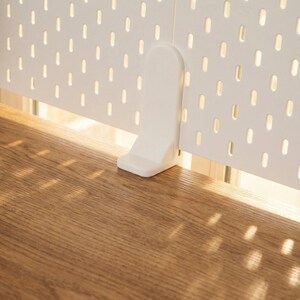 May include: A white pegboard panel with evenly spaced oval holes, supported by a white bracket. The pegboard is positioned above a wooden surface, with sunlight casting shadows.