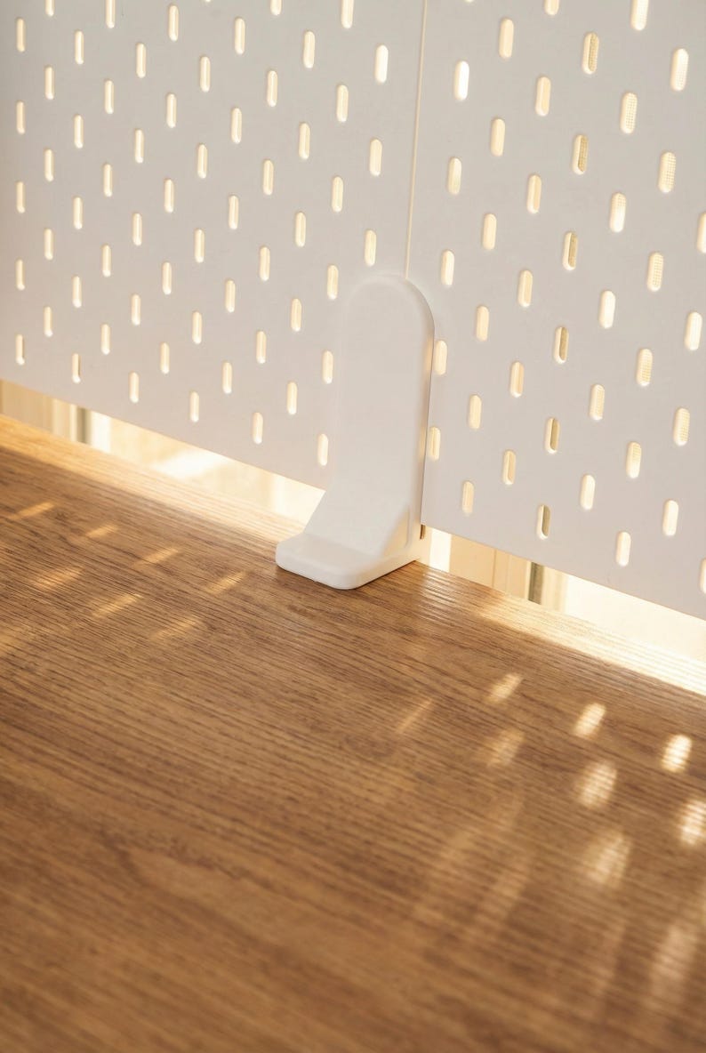 May include: A white pegboard panel with evenly spaced oval holes, supported by a white bracket. The pegboard is positioned above a wooden surface, with sunlight casting shadows.