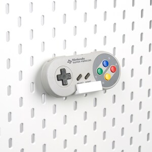 May include: A light gray Nintendo Super Famicom video game controller featuring a dark gray directional pad, two small gray buttons labeled Select and Start, and four colored action buttons labeled X, Y, A, and B, resting on a white wall mount.