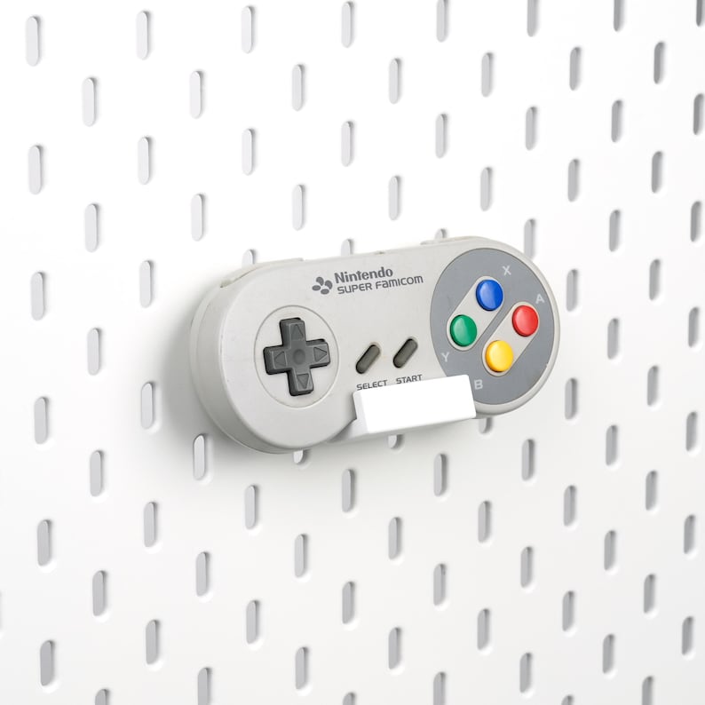 May include: A light gray Nintendo Super Famicom video game controller featuring a dark gray directional pad, two small gray buttons labeled Select and Start, and four colored action buttons labeled X, Y, A, and B, resting on a white wall mount.
