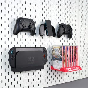 May include: A wall-mounted gaming setup featuring a black Nintendo Switch console, controllers, and game cases. The setup includes a charging dock, controller holders, and a red game case holder, all mounted on a white pegboard.