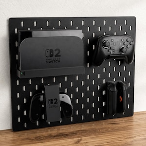 Peut inclure: Un organisateur de pegboard noir avec une station d'accueil Nintendo Switch, une manette et des Joy-Cons. Le pegboard comporte des trous espacés et est monté sur une surface en bois. Le logo Nintendo Switch est visible.