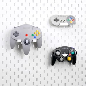 Puede incluir: Tres mandos de videojuegos: un mando gris de Nintendo 64, un mando gris de Super Nintendo y un mando negro de GameCube. Los mandos están montados sobre un fondo blanco de panel perforado.
