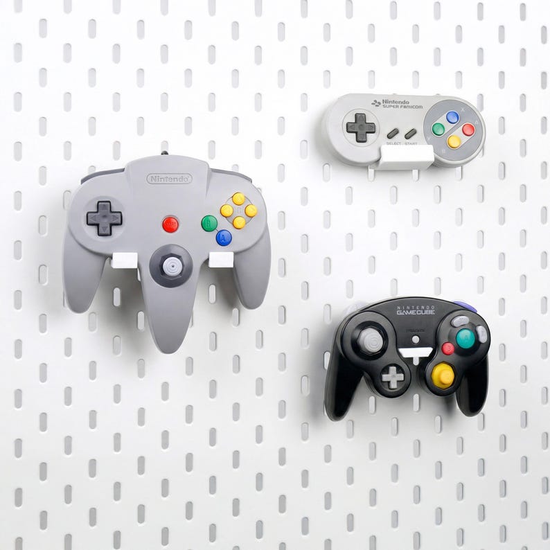 May include: A grey Nintendo 64 controller, a grey Super Famicom controller, and a black Nintendo GameCube controller mounted on a white pegboard. Each controller features its respective brand logo, directional pads, analog sticks, and colored buttons.