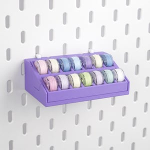 May include: A purple plastic organizer rack holding fourteen clear sewing machine bobbins wound with various pastel-colored threads, arranged in two rows of seven, mounted on a white pegboard with vertical oval slots.