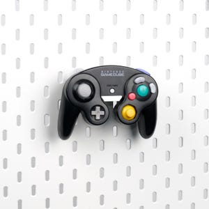 Puede incluir: Mando negro de Nintendo GameCube con botones de colores, incluyendo verde, rojo y amarillo. El mando tiene una cruceta, una palanca analógica y el logotipo de Nintendo GameCube. Se encuentra sobre un fondo blanco con un patrón de pequeños agujeros espaciados.