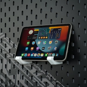 Puede incluir: Una tableta con una pantalla colorida está montada en un soporte blanco, sujeta a una panel perforado negro. La tableta muestra iconos de aplicaciones y una pantalla meteorológica. El panel perforado tiene un patrón de pequeños agujeros y un diseño geométrico.