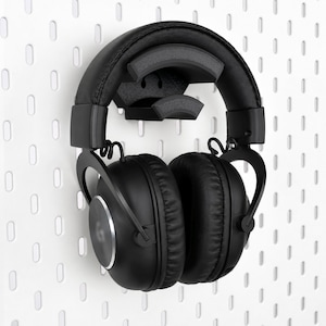 Puede incluir: Unos auriculares negros sobre un soporte de pared negro. Los auriculares cuentan con una diadema acolchada, almohadillas para los oídos y un logotipo circular en el lateral. El soporte está fijado a un panel perforado blanco con ranuras.