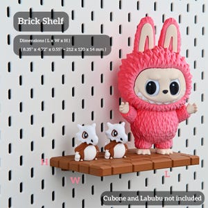 Puede incluir: Una estanter&iacute;a de ladrillo marr&oacute;n de 212 x 120 x 14 mm, que muestra una figura rosa y peluda de Labubu y dos figuras de Cubone. La estanter&iacute;a est&aacute; montada en una pared perforada blanca. El texto de la imagen dice "Brick Shelf" y "Cubone and Labubu not included."