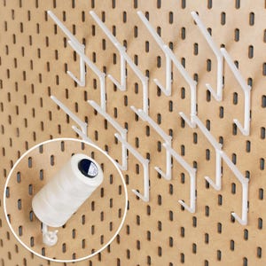 Puede incluir: Un conjunto de ganchos de plástico blanco para tablero perforado dispuestos en cuadrícula sobre un tablero color canela. Un recuadro circular muestra un carrete de hilo blanco colgado de uno de estos ganchos, con una etiqueta azul oscuro.
