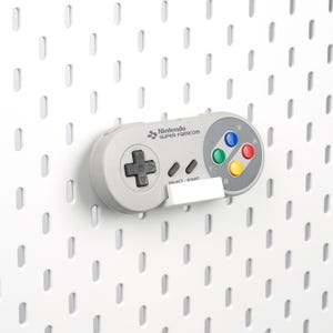 May include: A gray Nintendo Super Famicom controller mounted on a white pegboard. The controller has a directional pad, colored buttons, and the text "SELECT START". The background is a white pegboard with evenly spaced holes.