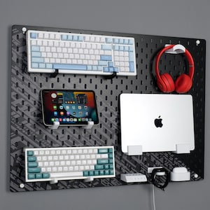 Puede incluir: Un panel perforado negro con varios accesorios tecnológicos. Incluye dos teclados, una tableta, un portátil, auriculares rojos, un reloj inteligente y una estación de carga. Los teclados tienen teclas blancas y azules o turquesas.