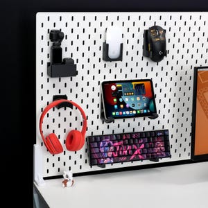 May include: A white pegboard with various tech accessories. Items include a red headset, a tablet, a keyboard, a mouse, and a camera. The accessories are mounted on the board with black holders. A small figurine sits on the desk.