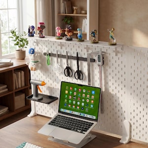 May include: A home office setup featuring a laptop on a stand, a white pegboard with various tools and figurines, and a wooden desk. The laptop screen displays colorful app icons. A glue gun and plush toy are also visible.