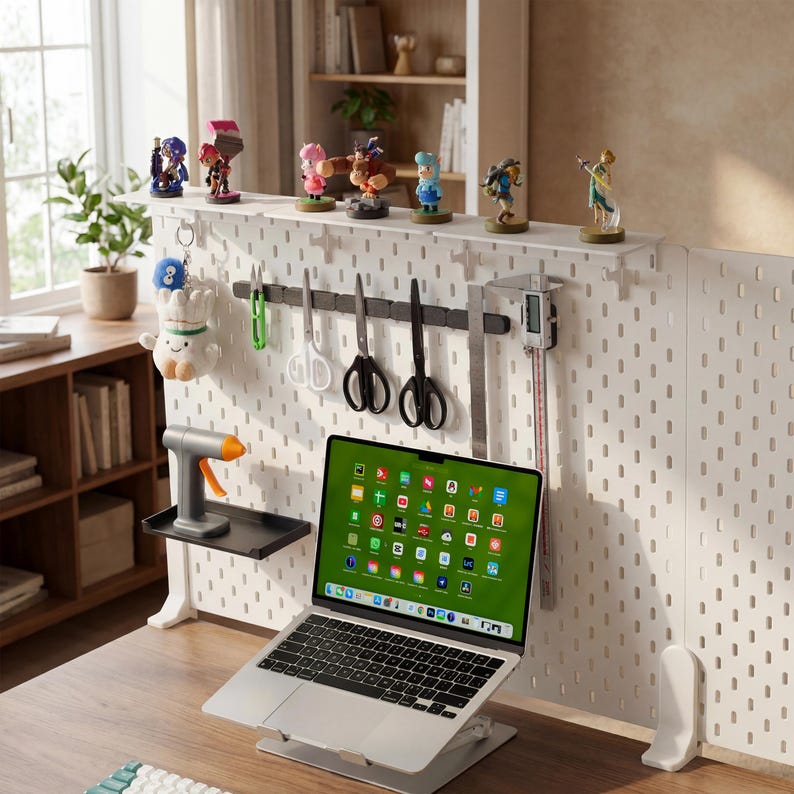 May include: A home office setup featuring a laptop on a stand, a white pegboard with various tools and figurines, and a wooden desk. The laptop screen displays colorful app icons. A glue gun and plush toy are also visible.