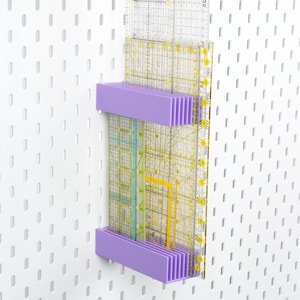 Puede incluir: Un organizador de pl&aacute;stico color lavanda montado en un tablero perforado blanco, dise&ntilde;ado para sostener varias reglas de acolchado de acr&iacute;lico transparente. El estante cuenta con dos barras horizontales con ranuras verticales.