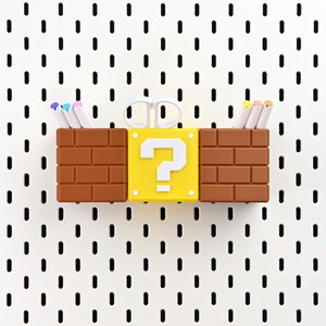May include: A novelty desk organiser featuring a yellow question mark block flanked by two brown brick-style containers. White scissors rest atop the question mark block, and colourful pens are in the containers. The organiser is set against a white pegboard.