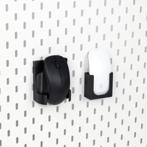 Puede incluir: Dos soportes negros para ratones de ordenador montados en una pared perforada blanca. Un soporte contiene un ratón negro y el otro un ratón blanco con el logotipo de Apple. Los soportes están diseñados para sujetar los ratones de forma segura.