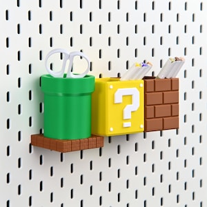 May include: A set of three colourful desk organisers in the style of a video game. The first is a green pipe, the second is a yellow question mark box, and the third is a brown brick block. White scissors and pens are inside.