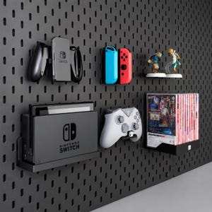 May include: A black pegboard wall organizer holding a Nintendo Switch console in its dock, a gray wireless controller, a pair of Joy-Con controllers, two small character figurines on a shelf, and a row of game cases in a black wall mount.