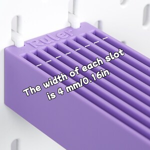 Puede incluir: Un organizador de pl&aacute;stico morado con m&uacute;ltiples ranuras paralelas, montado en un tablero perforado blanco. La palabra Ruler est&aacute; grabada en el borde superior. El texto indica: The width of each slot is 4 mm/0.16in, con flechas de medida.