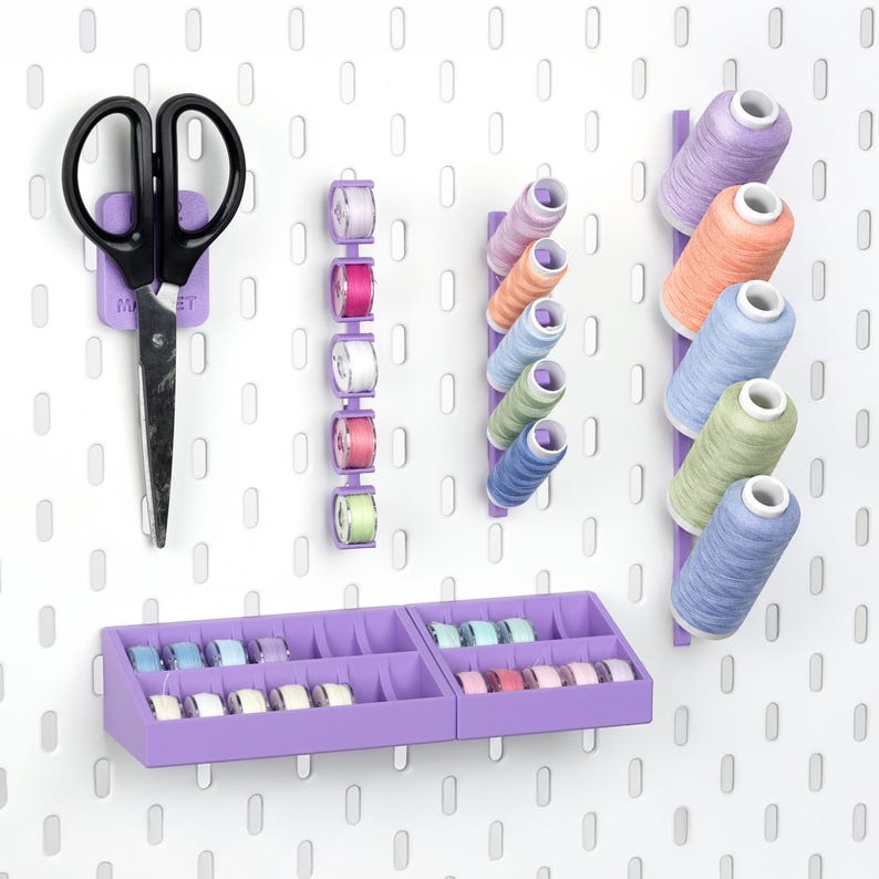 Puede incluir: Un organizador de pared perforado blanco con accesorios de pl&aacute;stico lavanda que incluyen un soporte para tijeras, soportes verticales para carretes de hilo de colores y dos bandejas escalonadas con varias bobinas de costura.