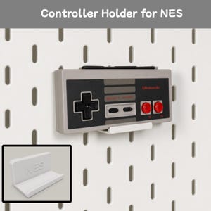 May include: A white L-shaped plastic controller holder for NES with the text NES embossed on the front. The holder is mounted on a white pegboard and supports a rectangular gray and black game controller featuring a black D-pad and two red buttons.