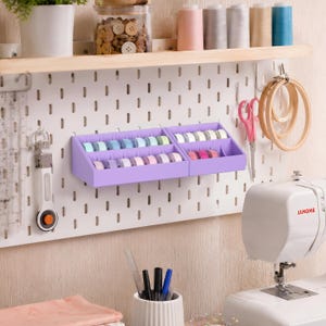 May include: A craft room scene with a white pegboard, displaying a purple thread organiser filled with colourful spools. Other items include sewing supplies, scissors, embroidery hoops, and a sewing machine. A wooden shelf holds jars of buttons and spools of thread.