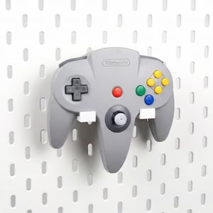 May include: A gray Nintendo 64 video game controller mounted on a white pegboard wall. The controller features a black directional pad, a central red start button, a gray analog stick, and four colored action buttons in yellow, green, and blue.