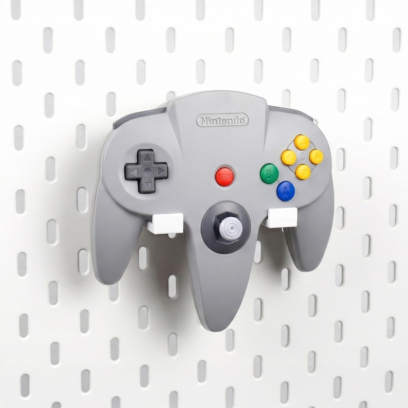 May include: A gray Nintendo 64 video game controller mounted on a white pegboard wall. The controller features a black directional pad, a central red start button, a gray analog stick, and four colored action buttons in yellow, green, and blue.