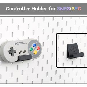 May include: A black plastic controller holder for SNES/SFC mounted on a white pegboard. The holder supports a grey Super Famicom controller featuring a black directional pad, grey select and start buttons, and four colored action buttons.