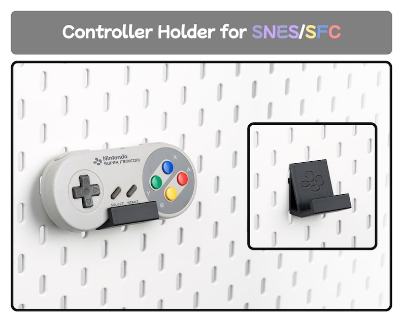 May include: A black plastic controller holder for SNES/SFC mounted on a white pegboard. The holder supports a grey Super Famicom controller featuring a black directional pad, grey select and start buttons, and four colored action buttons.
