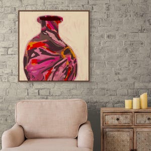 May include: Abstract painting of a vase in a wooden frame. The vase features a vibrant mix of pink, red, and yellow brushstrokes against a cream background. The artwork is displayed above a beige armchair and a wooden cabinet.