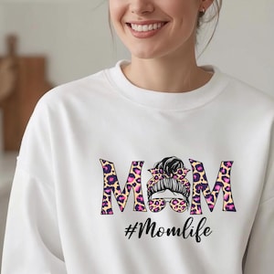 Può includere: Felpa bianca con la scritta "MOM" in stampa leopardata con accenti rosa. La "O" è una grafica di una donna con occhiali da sole e uno chignon. Il testo "#Momlife" è sotto la parola "MOM".