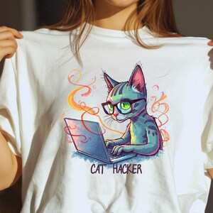 Può includere: T-shirt bianca con un gatto dei cartoni animati con gli occhiali che lavora su un laptop. Il gatto è blu e grigio con occhi verdi. Il testo "CAT HACKER" è sotto il gatto.