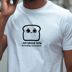 Può includere: T-shirt bianca con una fetta di pane a cartoni animati sorridente. Il testo "i AM bREAD NOW. birthdiay activated." è stampato sotto. La maglietta è realizzata in un materiale morbido.