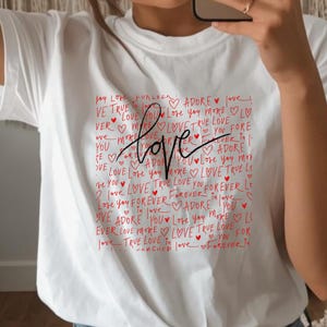 Può includere: T-shirt bianca con grafica "Love" rossa. Il design presenta la parola "Love" in corsivo, circondata da parole più piccole e forme di cuore. Uno stile casual e romantico.