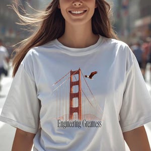Può includere: T-shirt bianca con il Golden Gate Bridge e un'aquila in volo sopra. Il testo "Engineering Greatness" è stampato sotto. La persona indossa un berretto da baseball rosso.