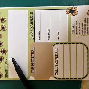 May include: ADHD daily planner notepad with a green and white design. Features sections for a loose schedule, top priorities, and call/text/email. Includes prompts for daily goals like moving the body and drinking water. A black pen rests on the notepad.