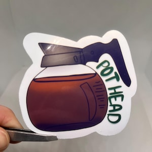 May include: A sticker featuring a cartoon coffee pot filled with a dark brown liquid. The handle and top are dark blue, and the words "POT HEAD" are in green. The sticker is held by tweezers.