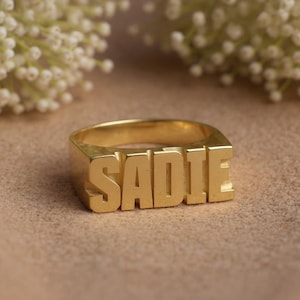 May include: A gold ring featuring the name "SADIE" in capital letters. The ring is rectangular and sits on a neutral surface, with white flowers in the background. This personalised ring is a statement piece of jewellery.