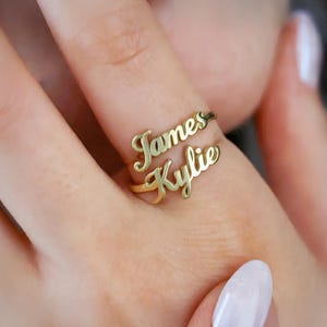 May include: A gold-colored double-band ring with the names "James" and "Kylie" in a cursive font. The ring is worn on a finger, showcasing the personalized design. The ring's elegant script and warm tone create a personalized jewelry piece.