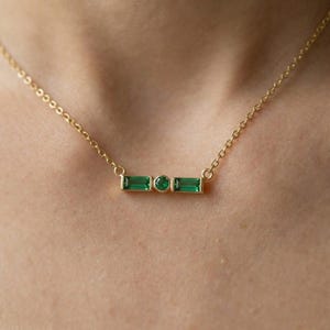 May include: A gold necklace with a bar pendant, featuring three green gemstones. Two rectangular emeralds are either side of a round emerald. The necklace is suspended from a fine gold chain and worn around the neck.