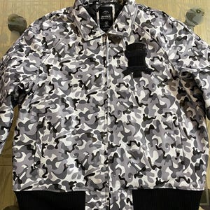 May include: A camouflage-patterned jacket in shades of gray, black, and white. The jacket features a black ribbed hem and cuffs, a zipper closure, and a black patch on the chest. The brand name "AVIREX" is visible on a label.