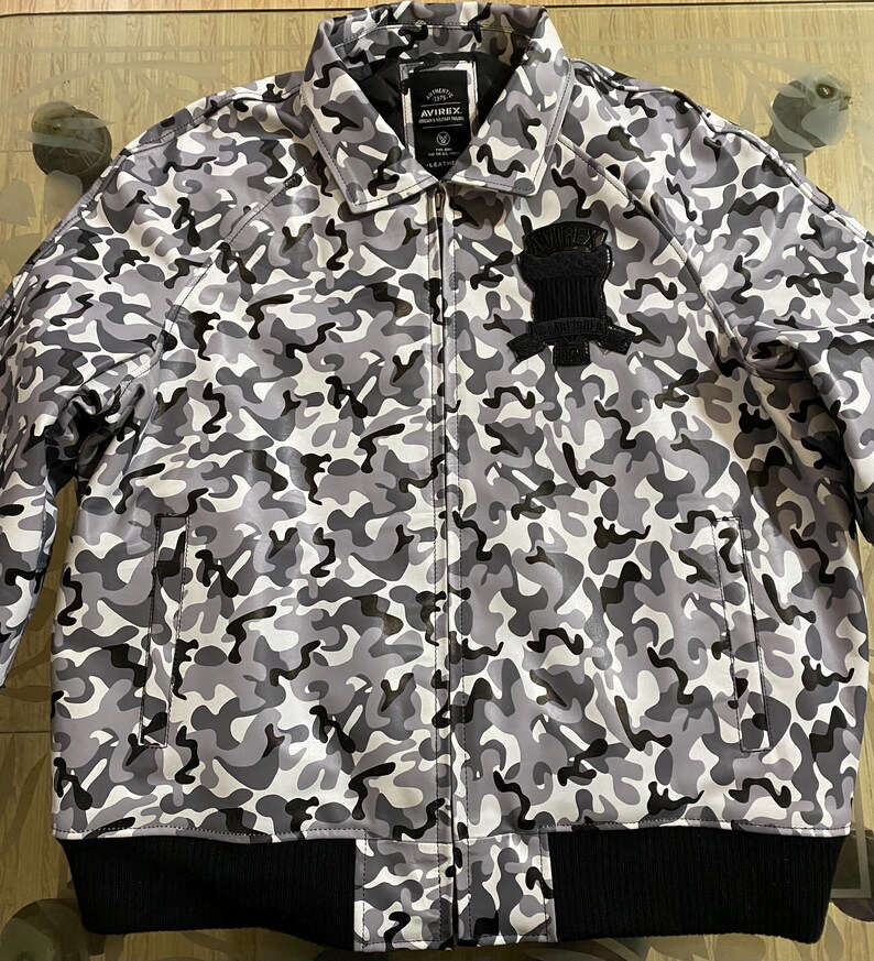 May include: A camouflage-patterned jacket in shades of gray, black, and white. The jacket features a black ribbed hem and cuffs, a zipper closure, and a black patch on the chest. The brand name "AVIREX" is visible on a label.