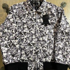 May include: A full-zip camouflage jacket in shades of gray and black. The jacket features a black ribbed hem and cuffs, a collar, and a patch on the chest. The jacket has the brand name "AVIREX" on the label.
