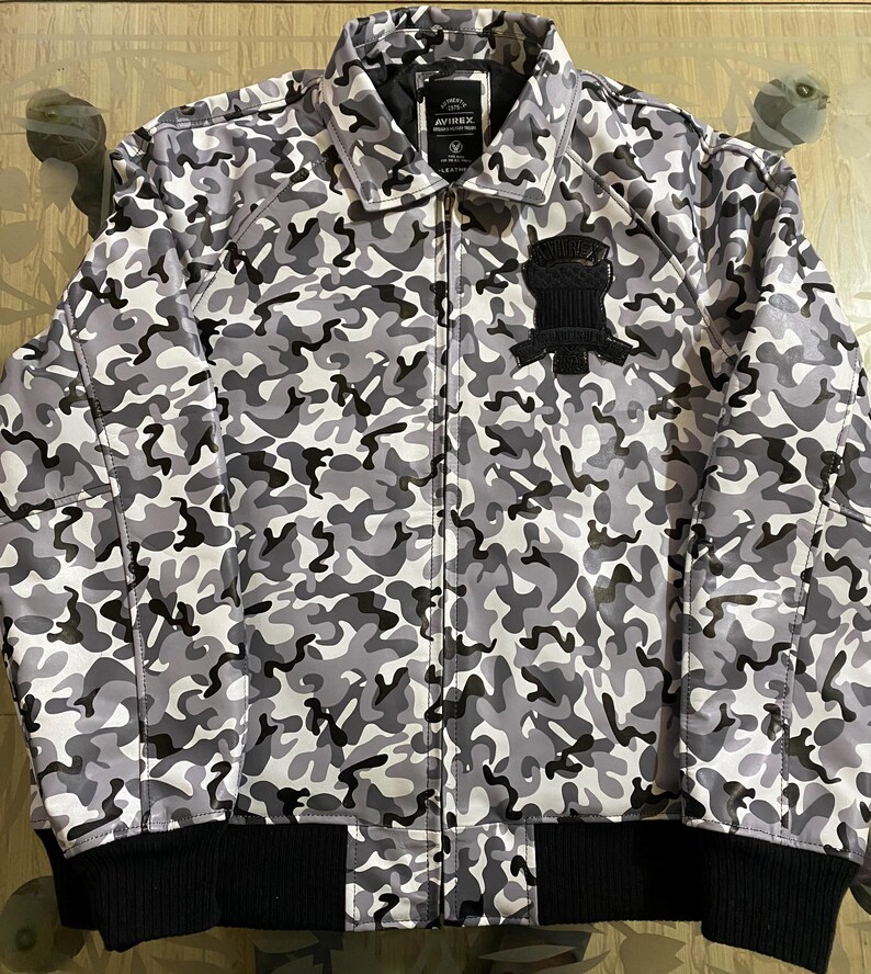 May include: A full-zip camouflage jacket in shades of gray and black. The jacket features a black ribbed hem and cuffs, a collar, and a patch on the chest. The jacket has the brand name "AVIREX" on the label.