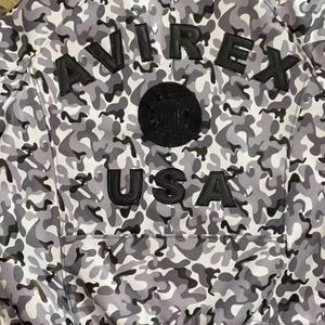 May include: A gray and white camouflage bomber jacket with the word "AVIREX" across the back in black lettering. Below the word "AVIREX" is a black circular patch and the word "USA" in black lettering. The jacket has a black ribbed hem.