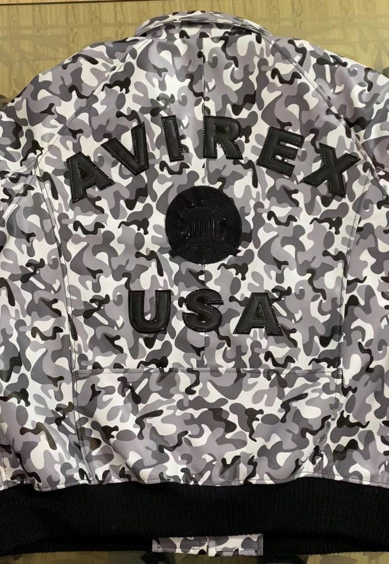May include: A gray and white camouflage bomber jacket with the word "AVIREX" across the back in black lettering. Below the word "AVIREX" is a black circular patch and the word "USA" in black lettering. The jacket has a black ribbed hem.