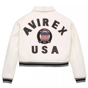 Puede incluir: Chaqueta color crema con ribetes a rayas blancas y negras. La parte trasera presenta la palabra "AVIREX" sobre un parche circular con un dise&ntilde;o de la bandera estadounidense y la palabra "USA" debajo. La chaqueta tiene un estilo bomber cl&aacute;sico.